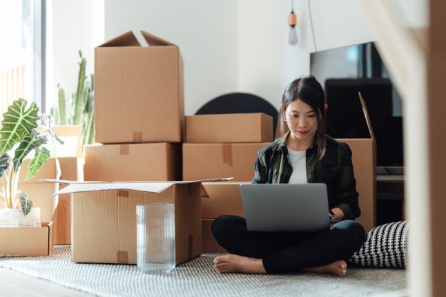 Young woman using laptop while sitting on the floor in new home with cardboard boxes.
